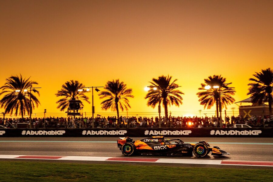 Závodní vozy Formule 1 na okruhu Yas Marina Circuit v Abú Dhabí při západu slunce nebo východu slunce, s palmami a luxusními budovami v pozadí.