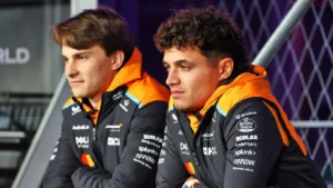 Dva závodníci v týmových bundách sedí vedle sebe v paddocku a soustředěně sledují dění na trati